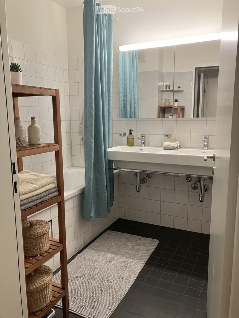 Modern bathroom with white tiles, double sink, bathtub, wooden shelf with towels and decorative items, and a blue shower curtain.