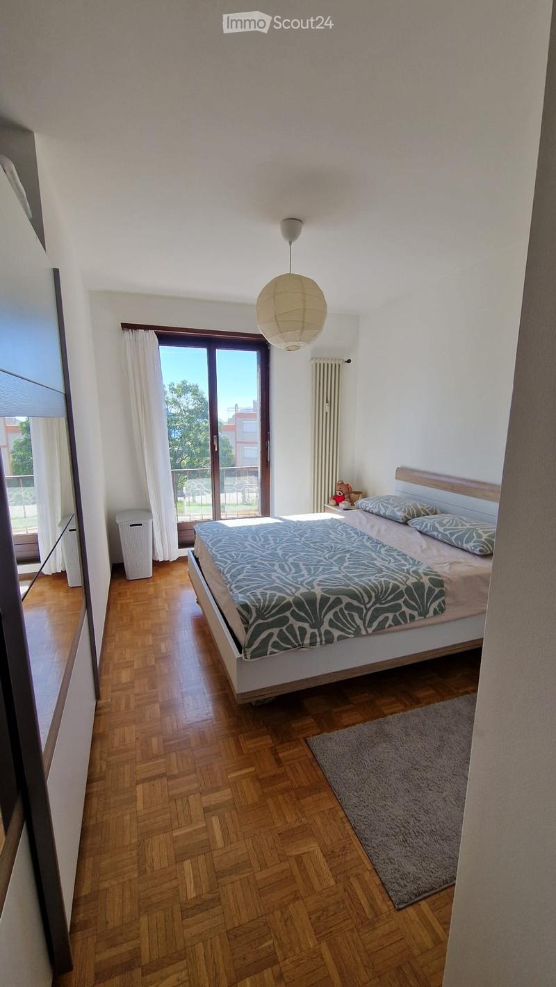 Bright bedroom with wooden flooring, large window with white curtains, bed with patterned duvet, nightstand with a toy, and a hanging paper lamp.