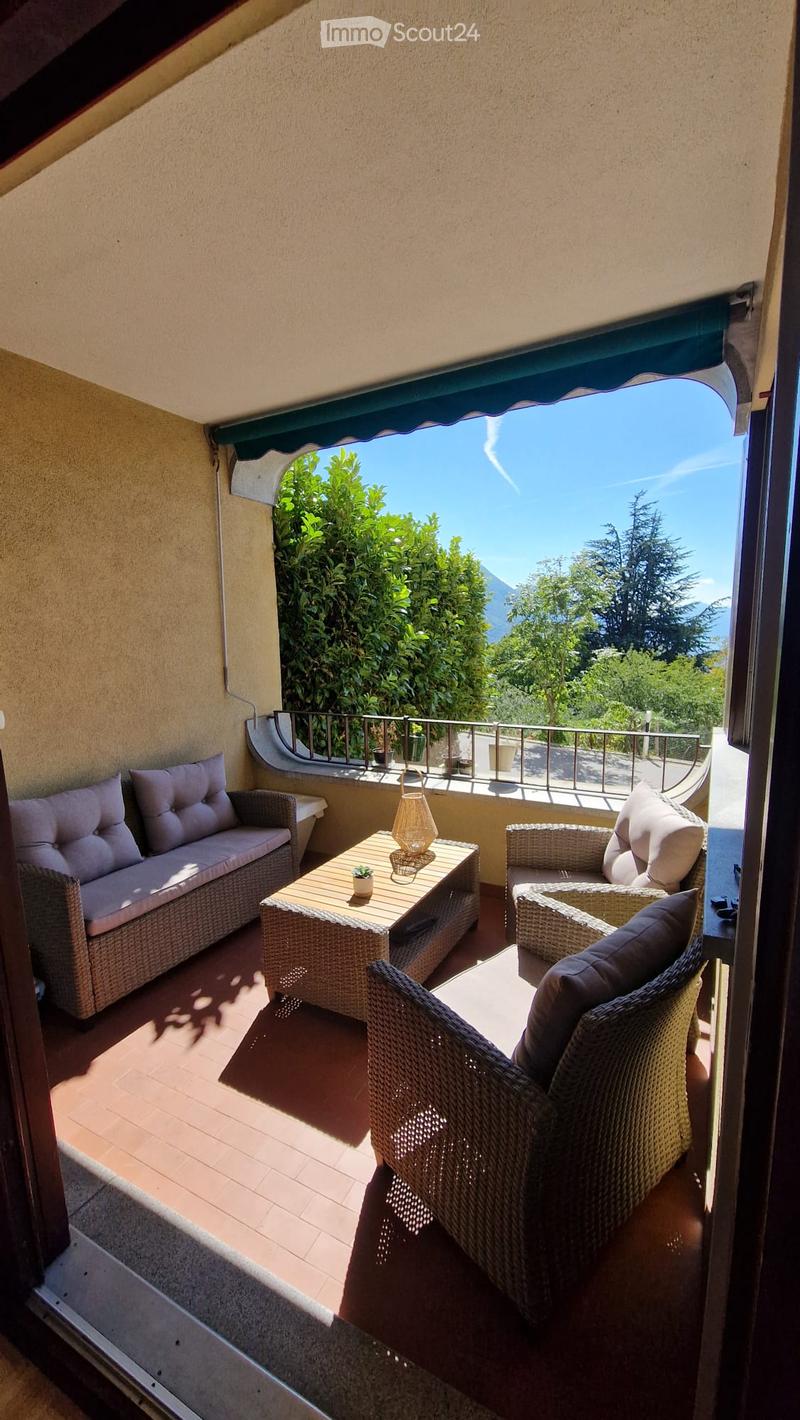 Balcony with wicker furniture, including a sofa, table, chairs, and a lamp, overlooking a scenic view of greenery and mountains.
