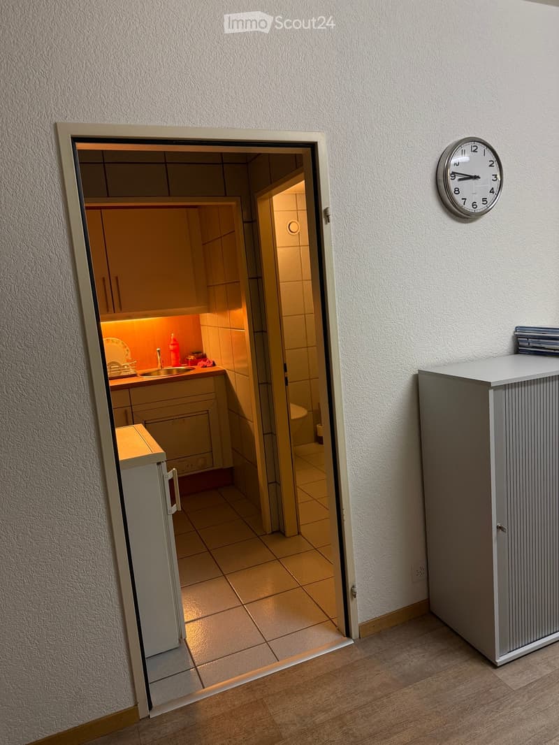 A narrow hallway with a wooden floor, leading to a small kitchen area with a sink, dishwasher, and cabinets. A clock is mounted on the wall to the right.