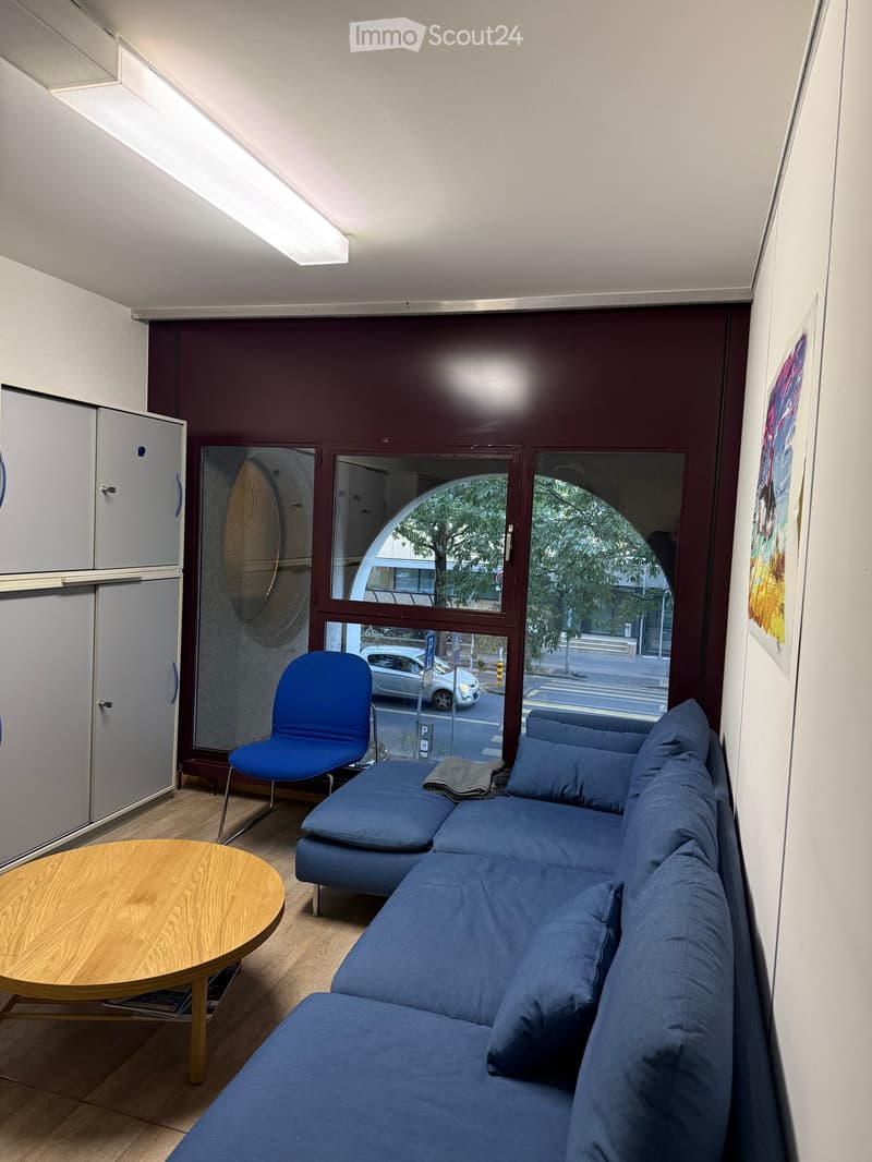 Modern living room with a blue sectional sofa, a blue chair, a round wooden table, and a white cabinet. Large windows offer a view of the street.