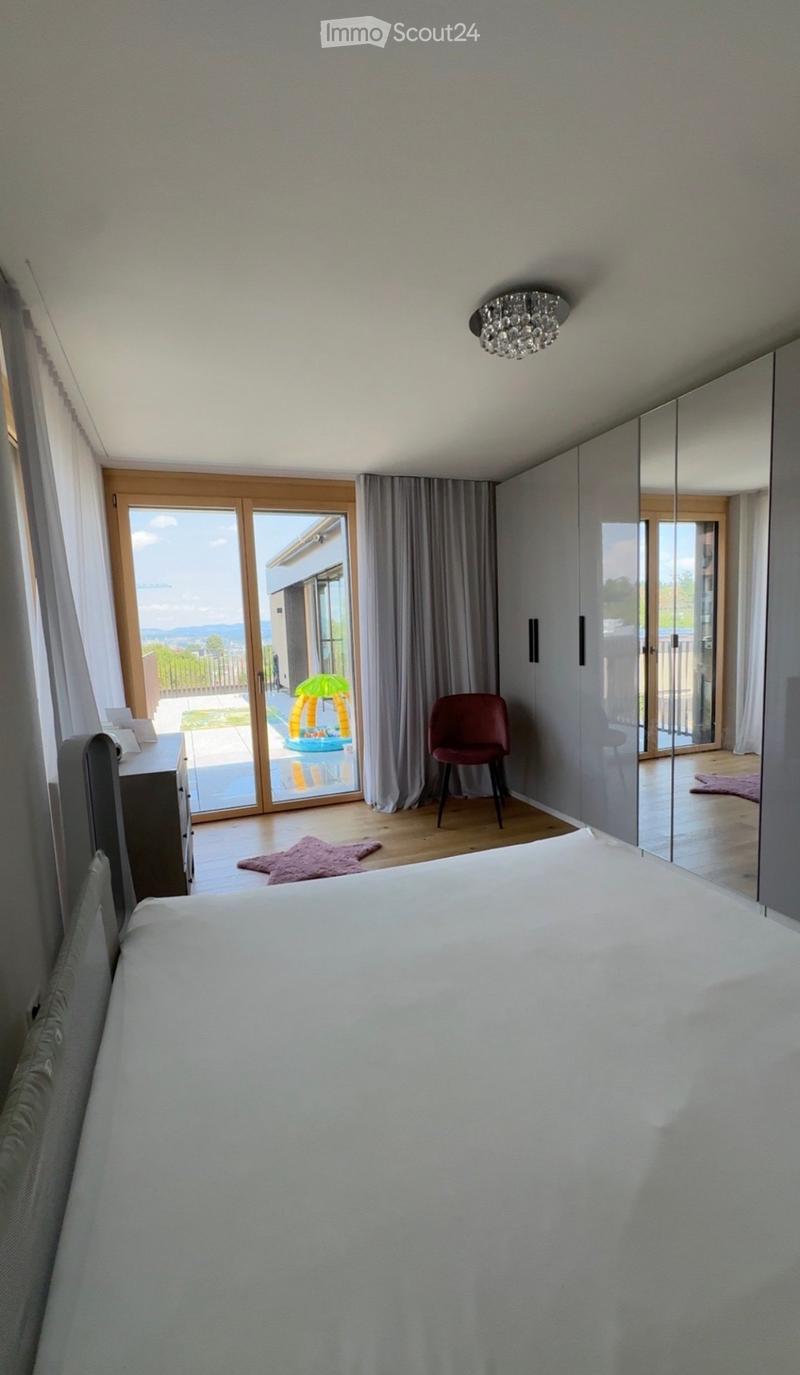 Modern bedroom with a large bed, a red chair, a mirror, and a view of a balcony through large glass doors.