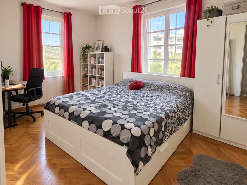 Bright bedroom with wooden flooring, large windows with red curtains, a white bed with a patterned duvet, a desk with a chair, a bookshelf, and a mirror.