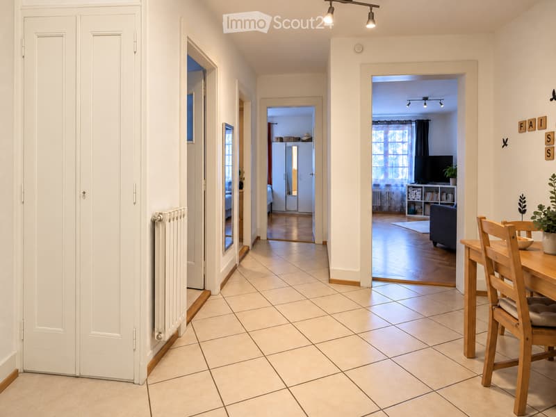 Bright hallway with tiled floor, white walls, and several doors leading to different rooms. Visible rooms include a living area with a sofa, TV, and dining table with chairs.