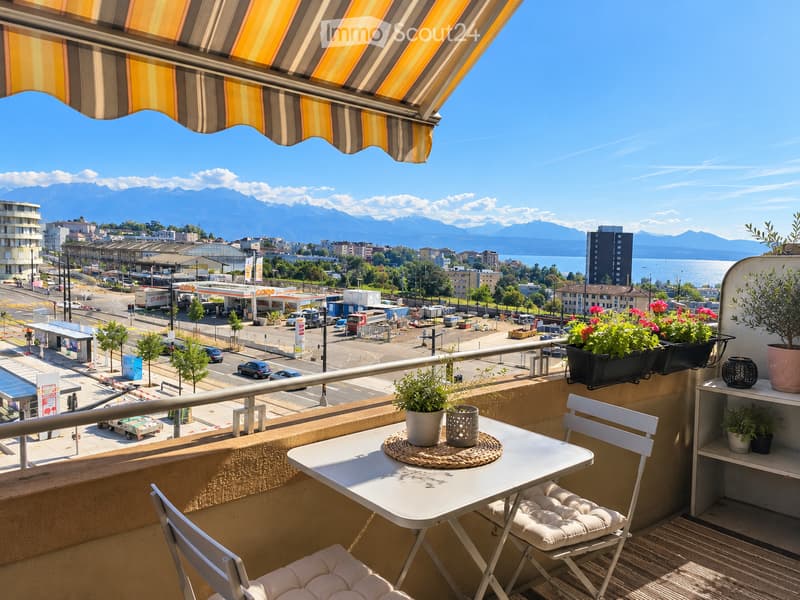 Balcony with a table, chairs, and potted plants, overlooking a cityscape with mountains in the background.