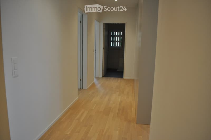 A clean, well-lit hallway with light wooden flooring and white walls. The corridor features several doors and a switch panel on the left side.