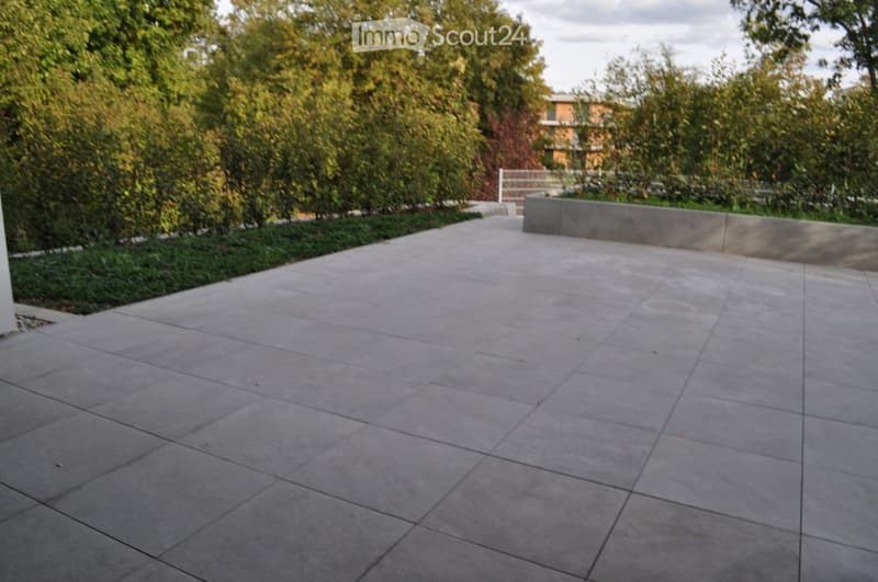 A paved outdoor area with large concrete tiles, surrounded by greenery and a low concrete wall on one side.