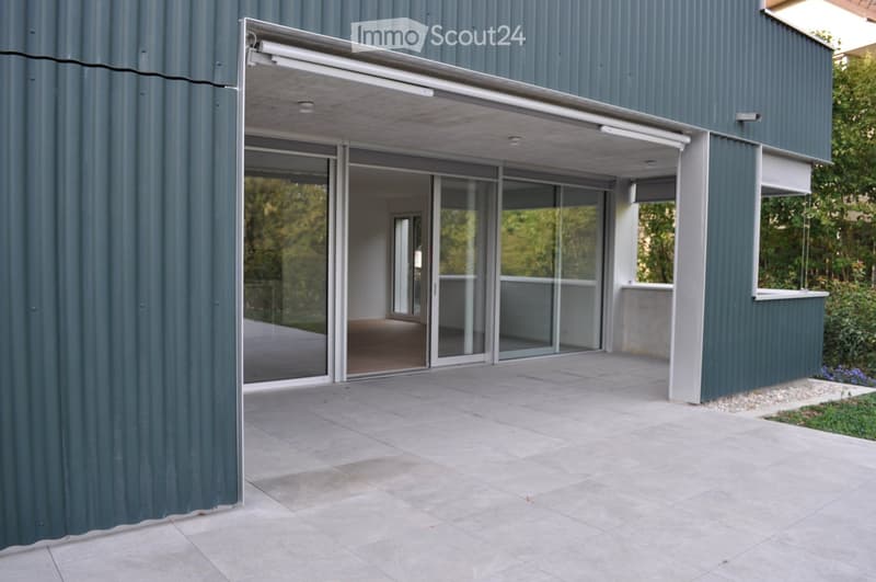 Modern building with large glass sliding doors leading to a covered outdoor area, grey tiled floor, and greenery visible through the glass.