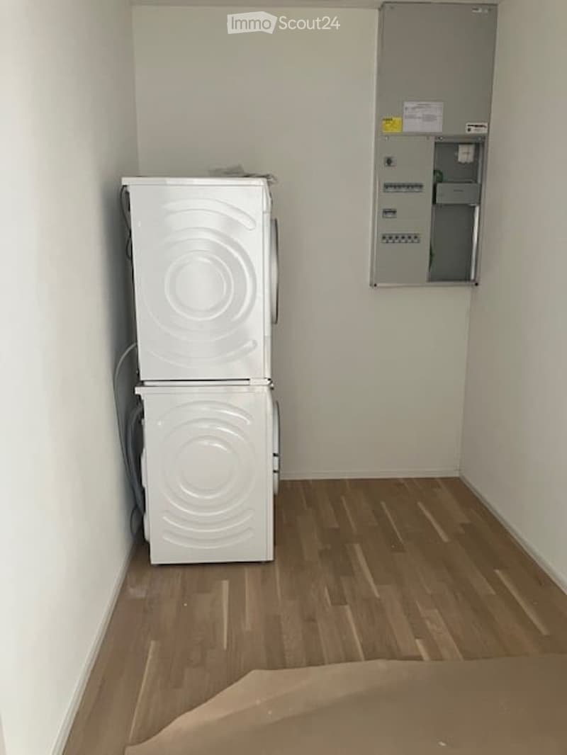 Empty room with wooden flooring, white walls, and a stacked washer/dryer unit in the corner. Electrical panel visible on the wall.