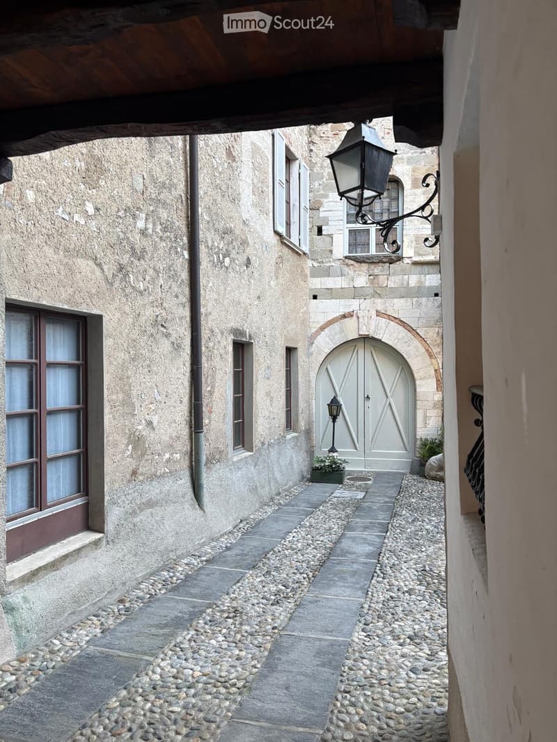 Narrow cobblestone alleyway between two stone buildings with arched doorways and windows, featuring classic street lamps and small potted plants.