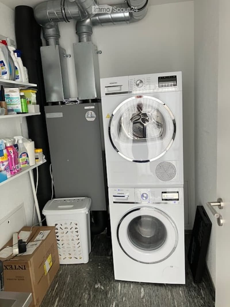 A compact laundry area with a stacked washing machine and dryer, a small refrigerator, shelves with cleaning supplies, and a laundry basket.