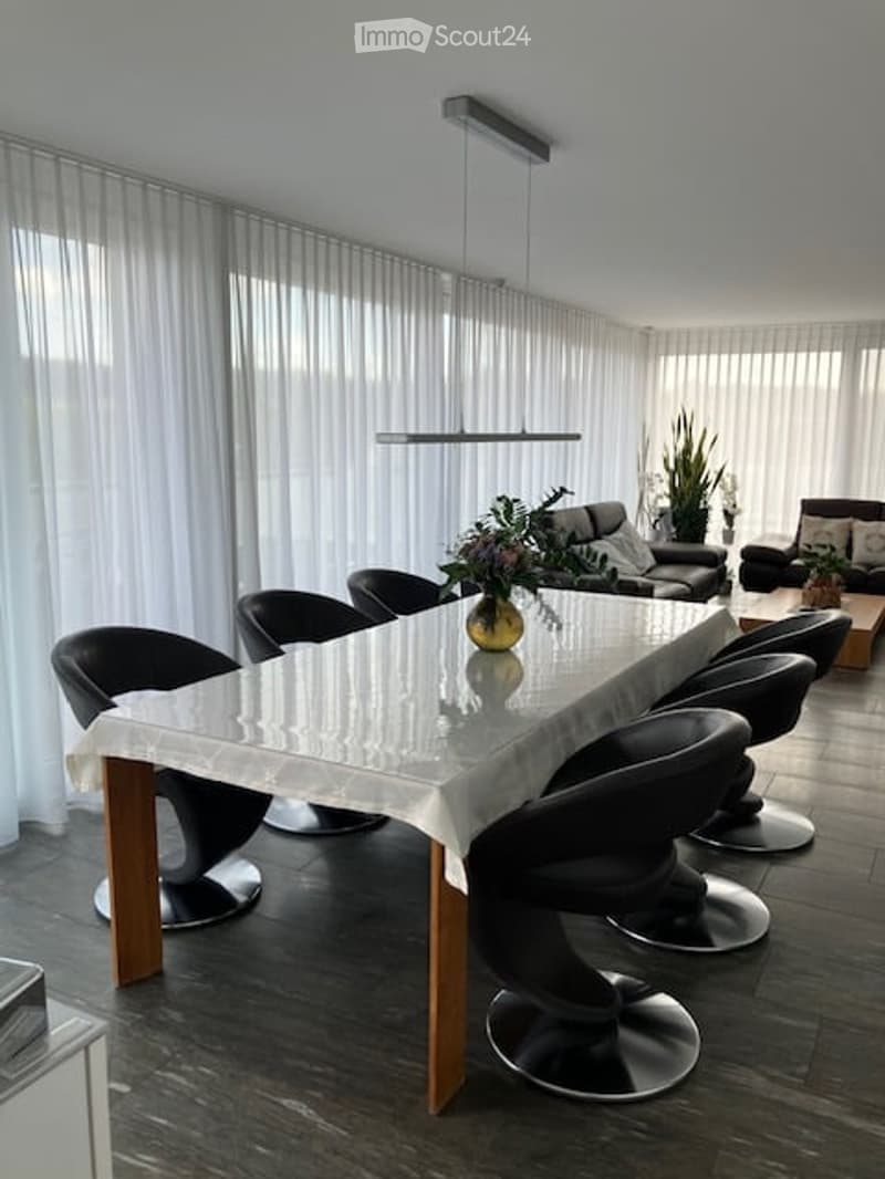 Modern dining area with a sleek glass dining table, black chairs, and a sofa in the background. Large windows with white curtains allow natural light to fill the space.