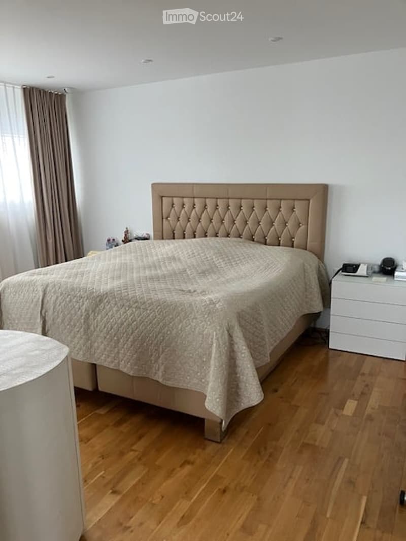 Modern bedroom with a large upholstered headboard, a neatly made bed, a nightstand, and a curtained window.