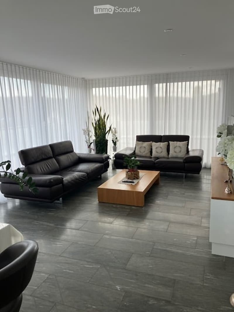 Modern living room with dark grey leather sofas, a wooden coffee table, and large windows with vertical blinds. The room features a dark tiled floor and decorative plants.