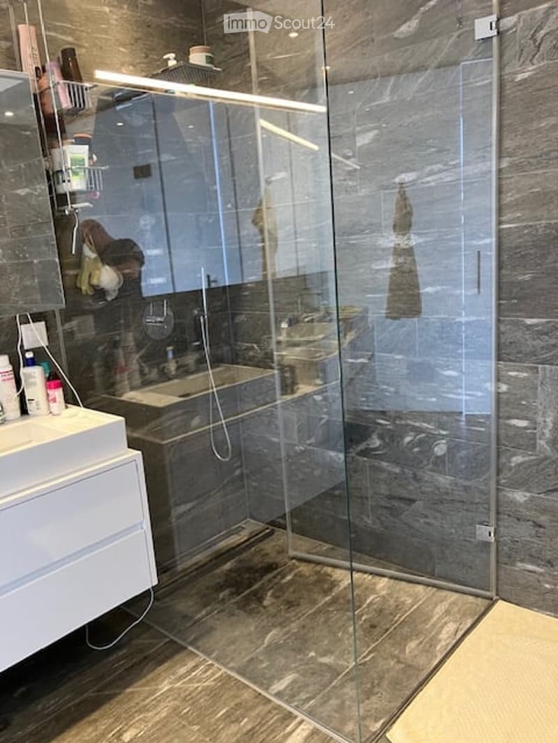 Modern bathroom with a large glass shower stall, a white sink with a mirror, and a tiled floor. The shower features a handheld showerhead and a built-in seat. A towel is visible on the wall.