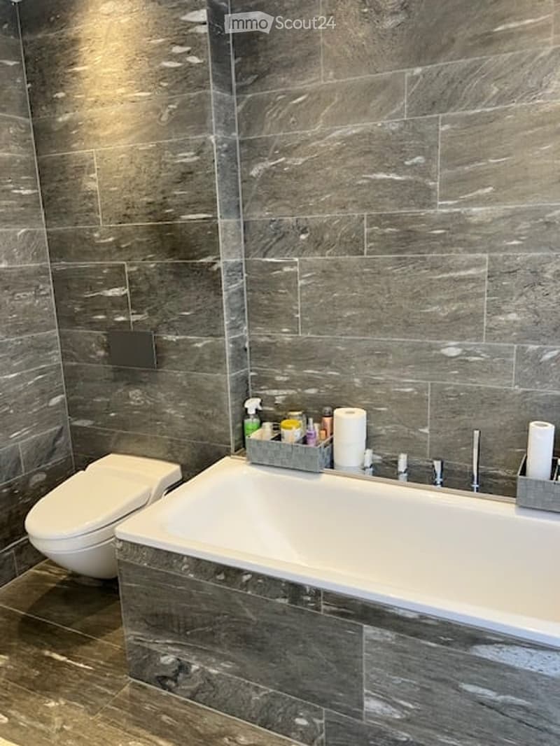 Modern bathroom with dark grey marble tiles, a large bathtub, toilet, and a tray with cleaning supplies and towels.