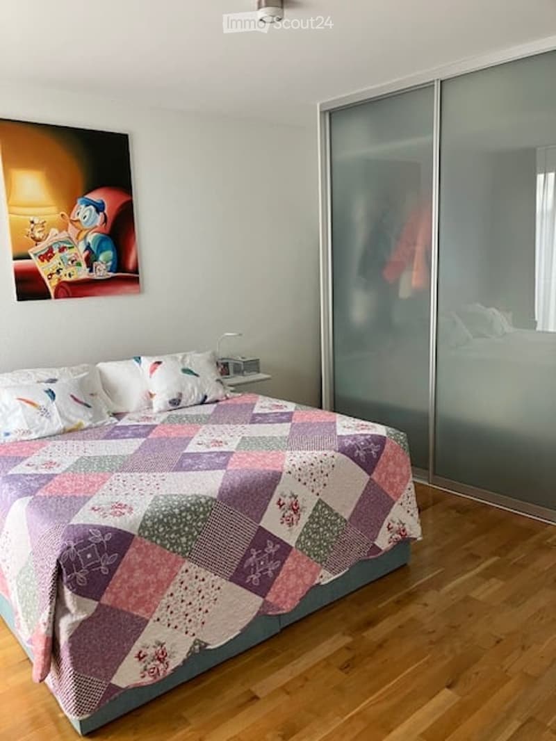 A well-lit bedroom with a wooden floor, a large bed with a colorful patchwork quilt, a nightstand with a lamp, a large mirror, and a picture frame on the wall.
