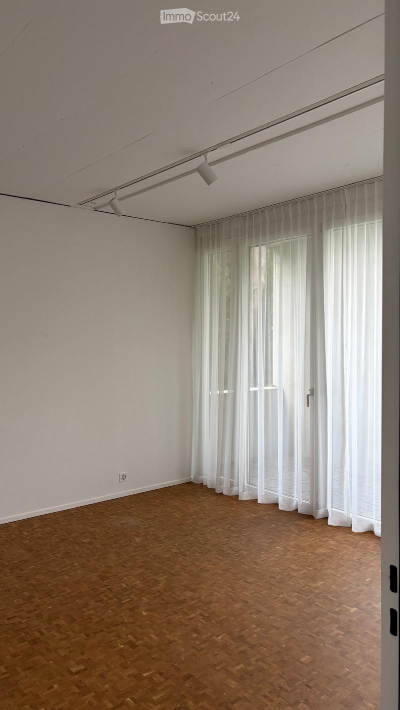 Empty room with wooden parquet flooring, white walls, large windows with sheer white curtains, and recessed ceiling lighting.