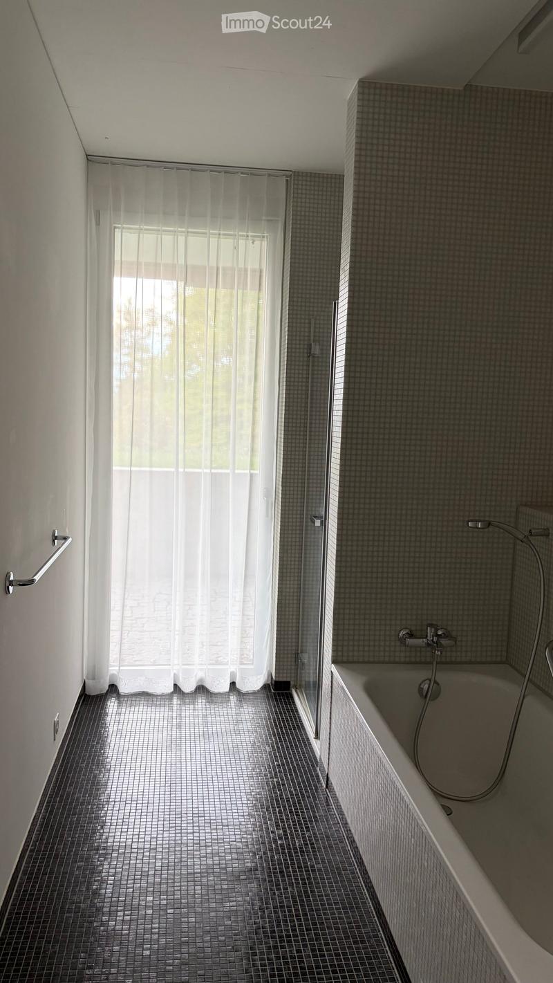 Modern bathroom with a bathtub, tiled walls and floor, and a large window with sheer curtains.