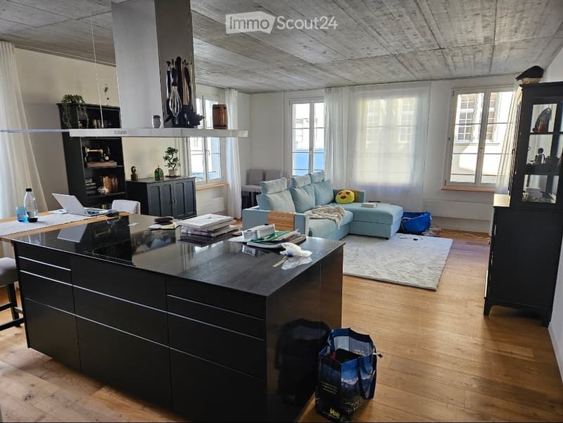 Open-plan living and dining area with a modern kitchen island, light blue sofa, wooden flooring, large windows, and various furniture and decor items.