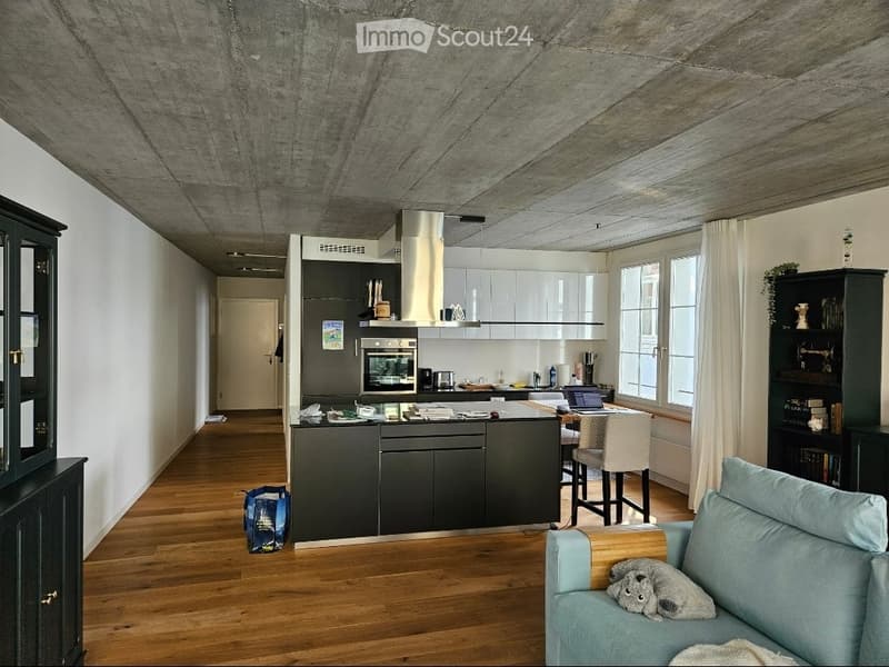 Modern open-plan living and kitchen area with wooden flooring, grey sofa, kitchen island, and large windows.