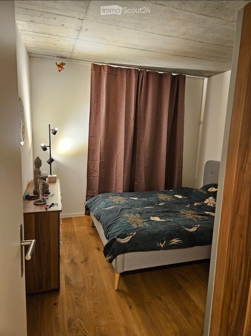 Cozy bedroom with wooden flooring, a bed with a patterned duvet, a nightstand with a lamp, a small table with decorative items, and brown curtains.