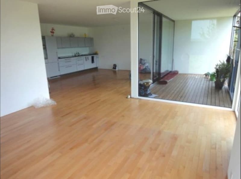 Empty room with light wooden flooring, white walls, and a large sliding glass door leading to a balcony area. The kitchen area is visible in the background with white cabinets and a countertop.