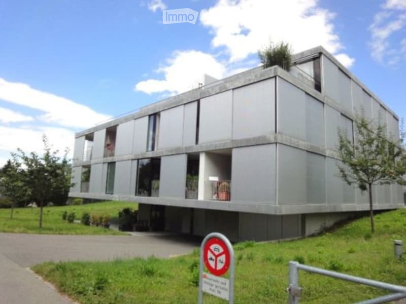 Modern, multi-story building with a sleek, grey concrete facade and large windows, elevated on stilts with a landscaped lawn and trees surrounding it.