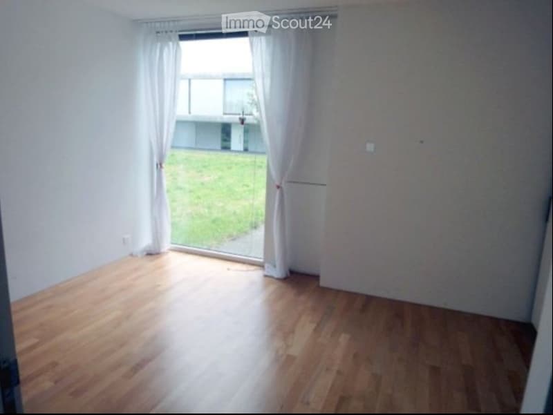 Empty room with wooden flooring, white walls, large window with white curtains, and a view of a green area outside.