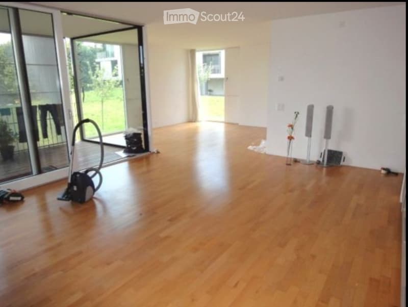Empty room with wooden flooring, large sliding glass doors leading to a balcony, and a vacuum cleaner placed near the entrance.