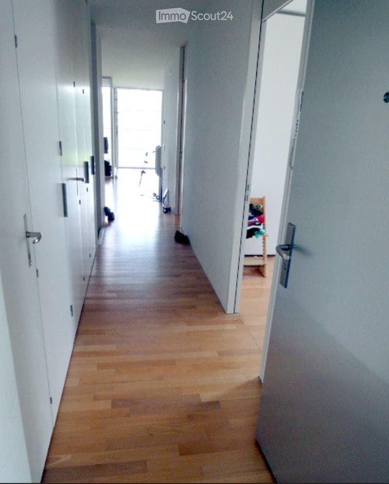 A long, narrow hallway with light wooden flooring and white walls. Doors line both sides, and the corridor leads to an open area at the end.