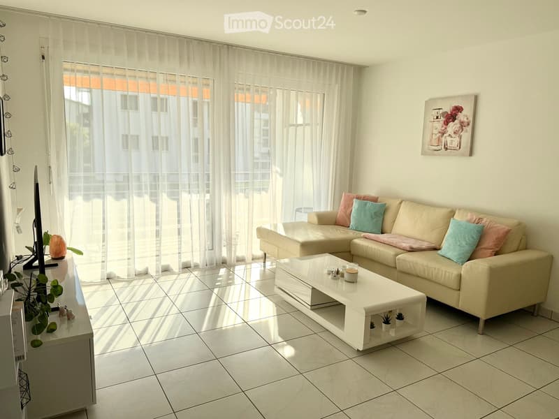 Modern living room with white tiled flooring, large windows with sheer curtains, a cream-colored sectional sofa with colorful cushions, a white coffee table, and a small plant on a side table.