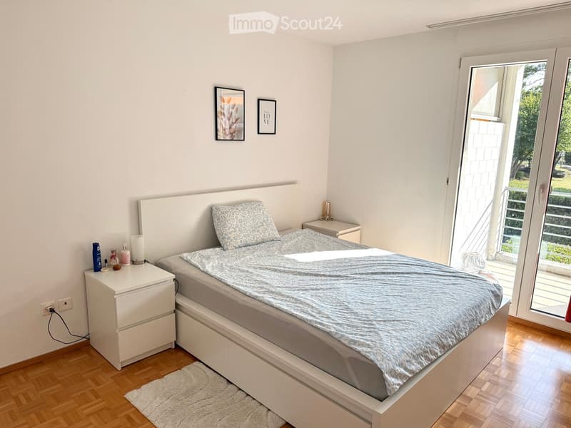 A modern bedroom with a large bed, nightstand, and picture frames on the wall. The room features wooden flooring and a sliding glass door leading to a balcony.