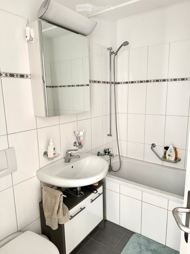 Modern bathroom with white tiles, a sink with a modern faucet, a bathtub with a handheld shower, a mirror cabinet, and a towel on a cabinet.