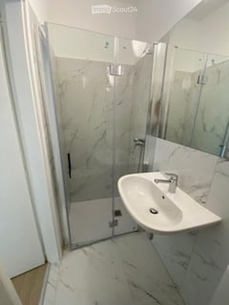 Modern bathroom with a white marble floor and walls, a glass-enclosed shower, a white sink with a chrome faucet, and a large mirror above the sink.