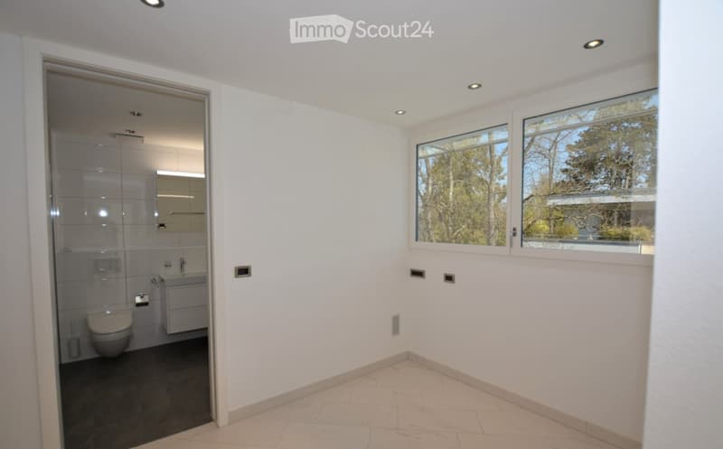 Modern bathroom with white tiled walls, a toilet, a sink, and a glass-enclosed shower. Large windows provide natural light and a view of greenery outside.