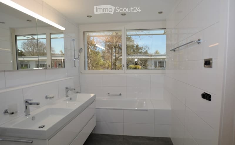 Modern bathroom with white tiles, double sink, bathtub, large mirror, and a window offering a view of trees and a building.