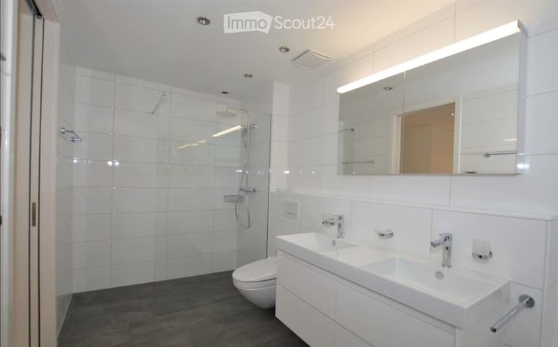 Modern bathroom with white tiles, a double sink, toilet, and a glass-enclosed shower area.