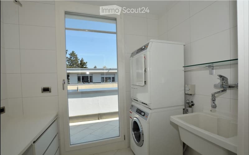 Modern bathroom with white tiles, a sink with a chrome faucet, a washing machine, and a large window offering a view of the outdoor area.