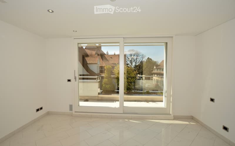 Empty room with white walls, marble tiled floor, and large sliding glass doors leading to a balcony with a view of neighboring houses and trees.