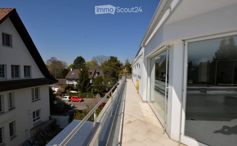 Balcony with modern glass railings, tiled floor, and large windows overlooking a residential neighborhood with houses, trees, and parked cars.