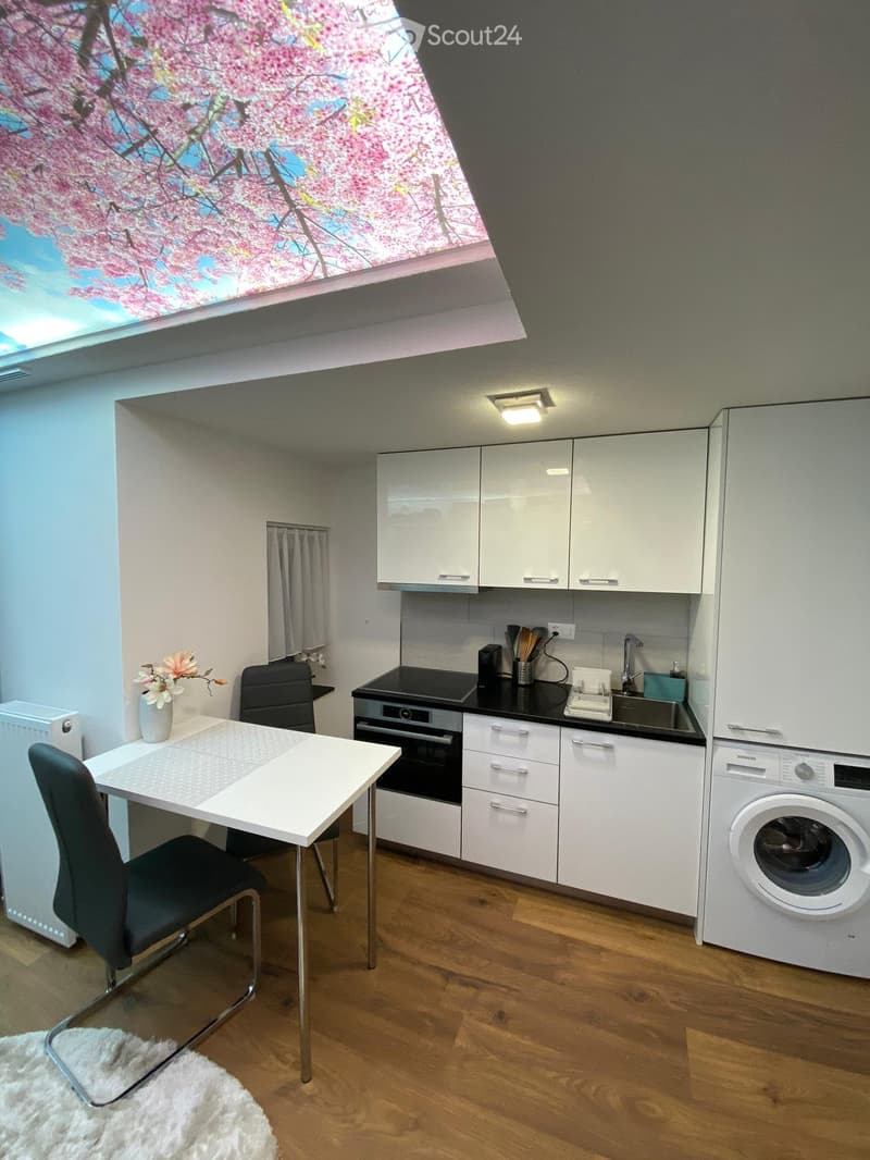 Modern kitchen with white cabinets, black countertop, built-in oven, sink, and washing machine. Includes a small dining table with chairs, a vase with flowers, and a radiator.