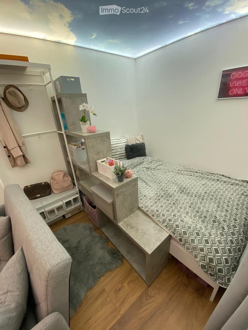 A cozy, compact room with a bed, sofa, small table, and shelves. The room features a wooden floor, a fur rug, and a ceiling with a sky-like design.
