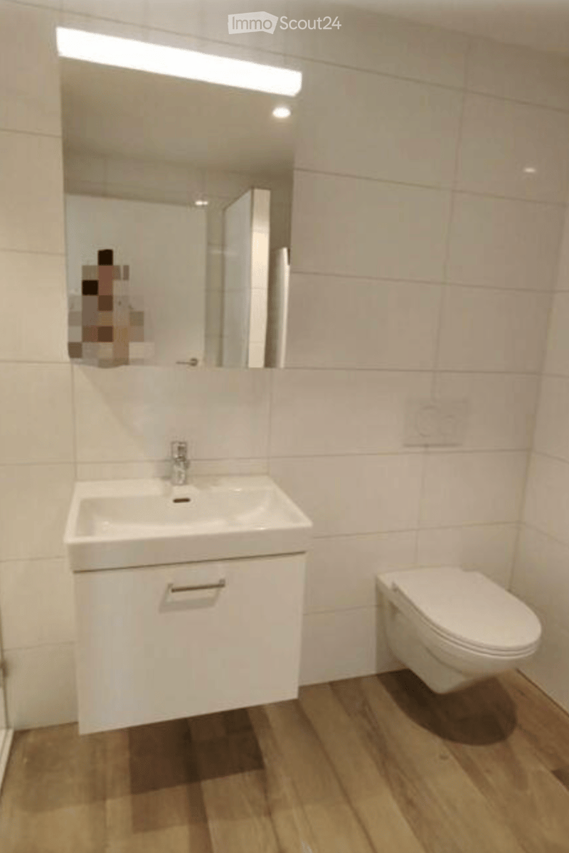 Modern bathroom with white tiles, a sink with a cabinet, a toilet, and a large mirror with built-in lighting.