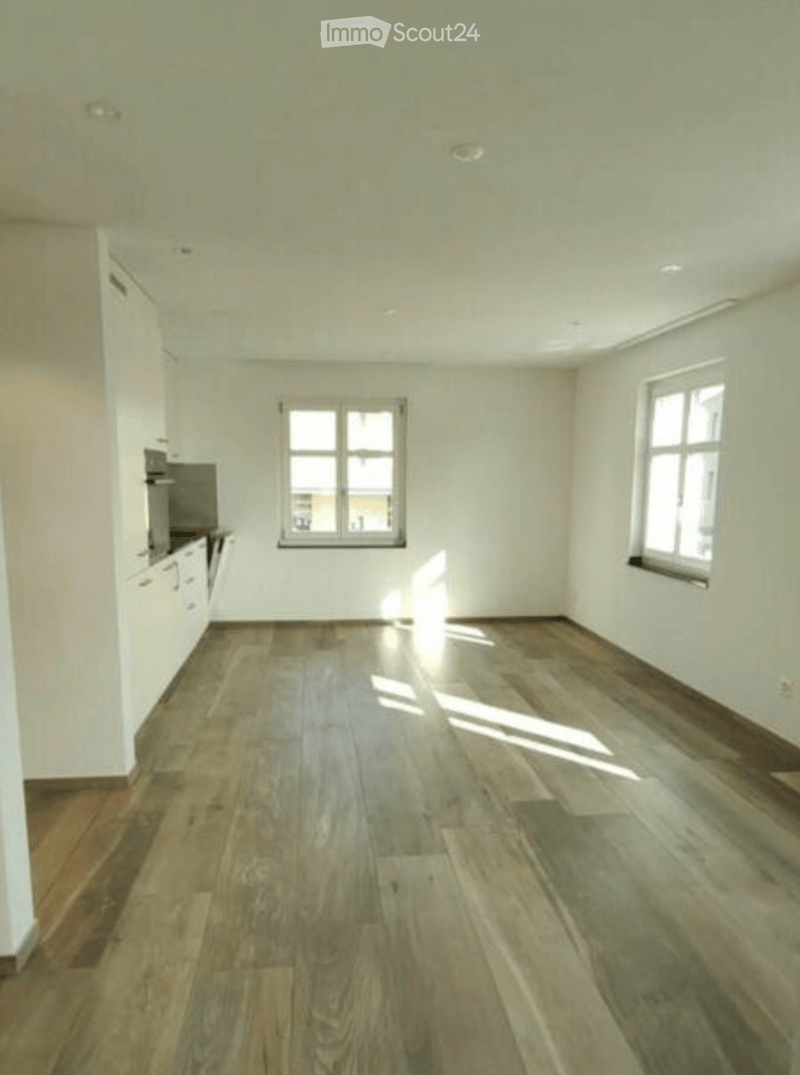 Empty room with wooden flooring, white walls, and two windows allowing natural light to enter. The room is unfurnished, showing only the basic structure and lighting fixtures.