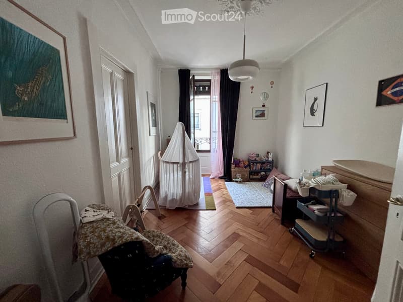 A cozy, well-lit room with wooden flooring, a baby cot, a small couch, a bookshelf, and a chair with a blanket draped over it.