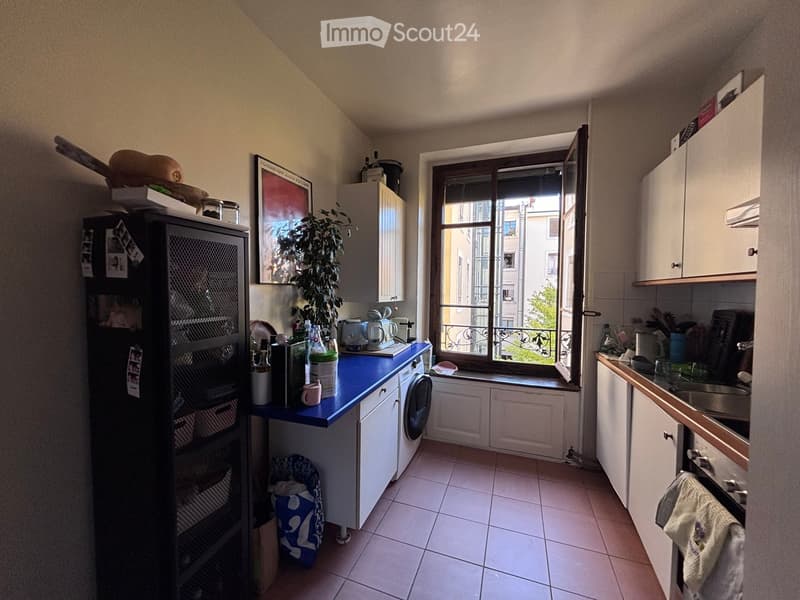 A compact kitchen with white cabinets, a blue countertop, a window overlooking a balcony, and various appliances including a refrigerator, oven, and sink.