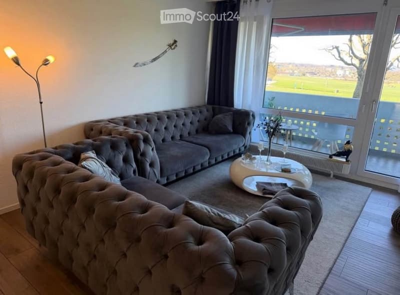 Modern living room with large windows offering a view, featuring a grey tufted sofa, a round coffee table, a floor lamp, and a small table with chairs near the window.