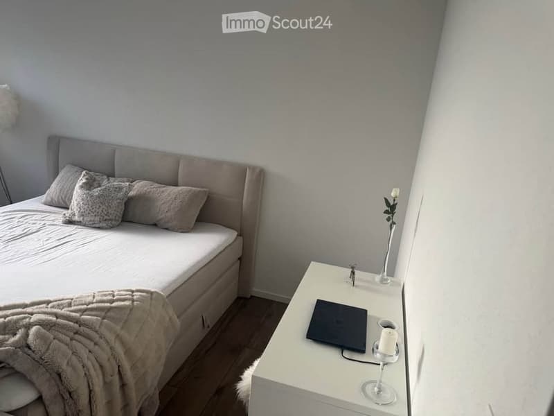 A modern bedroom with a large bed, nightstand, laptop, candle, and decorative vase with a flower. The room features light-colored walls and wooden flooring.