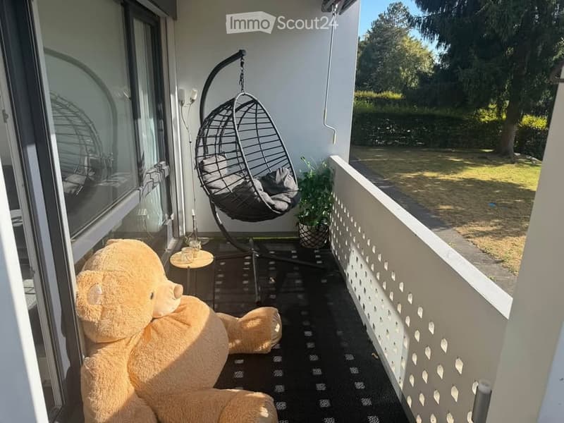 Balcony with a large teddy bear, a hanging wicker chair, a small table with a cup, and a potted plant. The balcony has a view of a green lawn and trees.